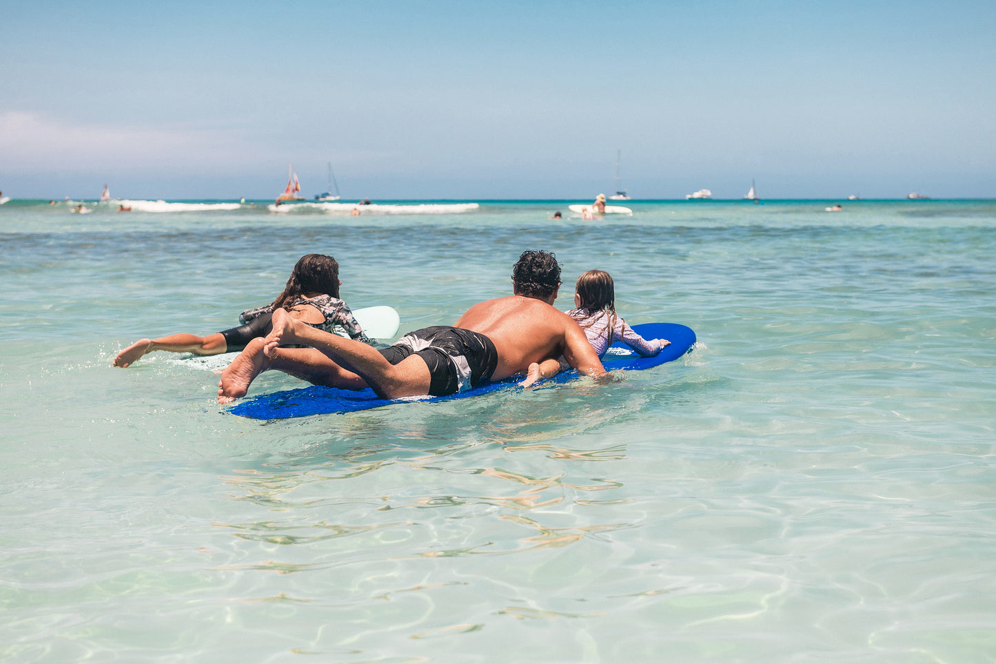 Two adults and a child lying on a blue Legion of Fun surfboard in clear ocean water with boats in the distance.