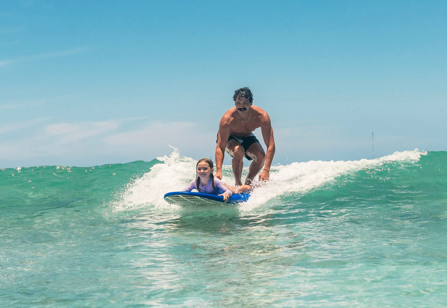 Father and daughter on a Legion of Fun surfboard riding a wave in clear blue water with a clear sky.