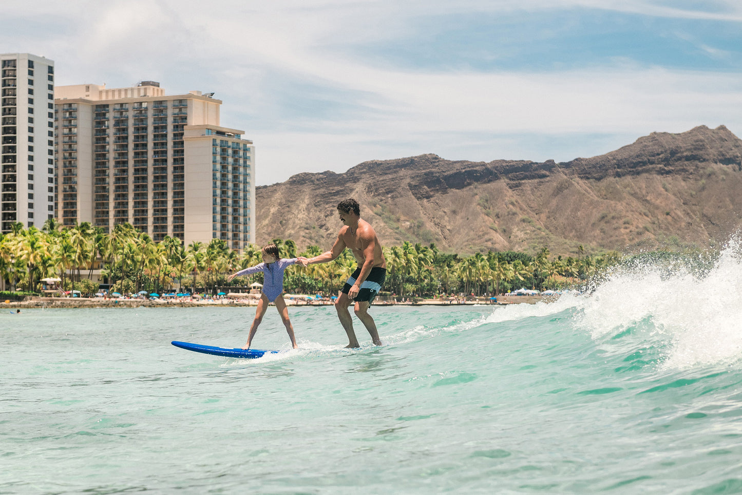 Father and daughter surfing a Legion of Fun board on a wave with a cityscape and mountains in the background