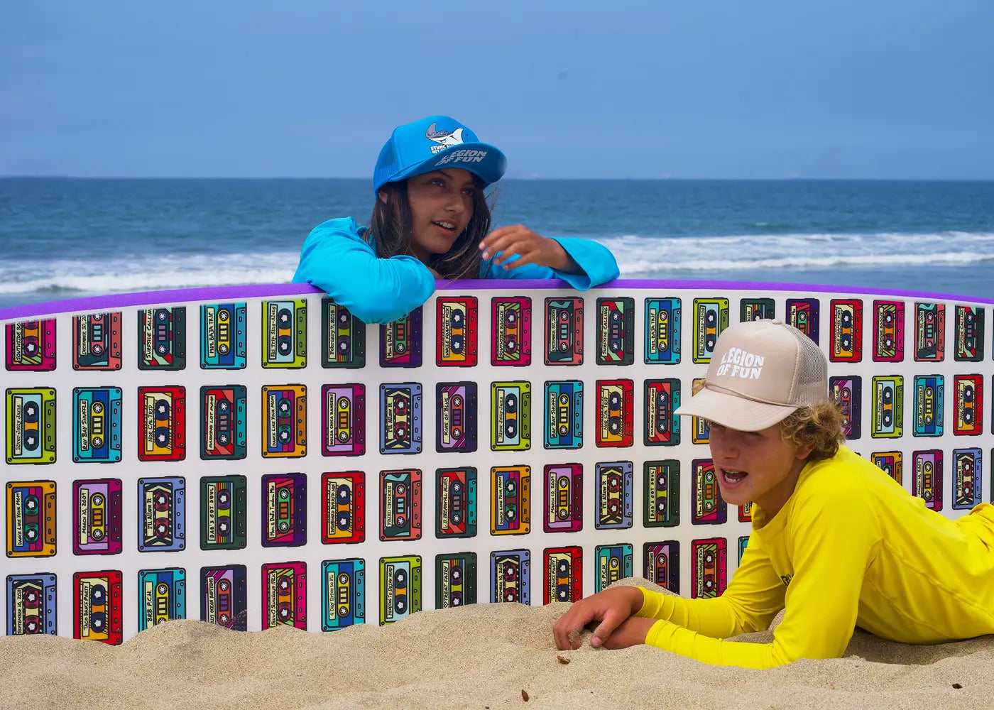 Two teens hang out on the sand in front of a Legion of Fun 10' Fun Wagon surfboard with mix tape design