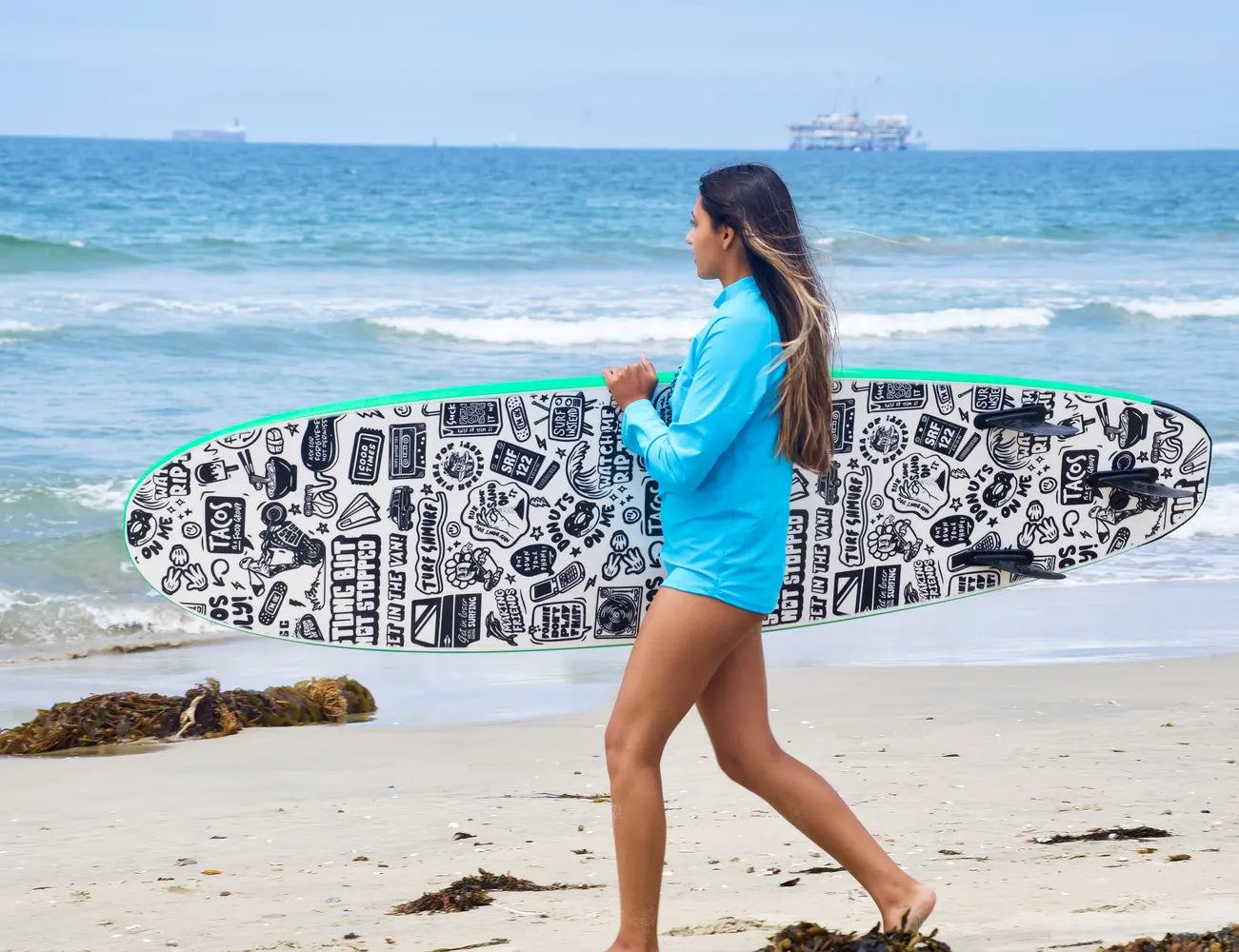 Teen girl with a blue rash guard holds a green foam surfboard heading for the ocean