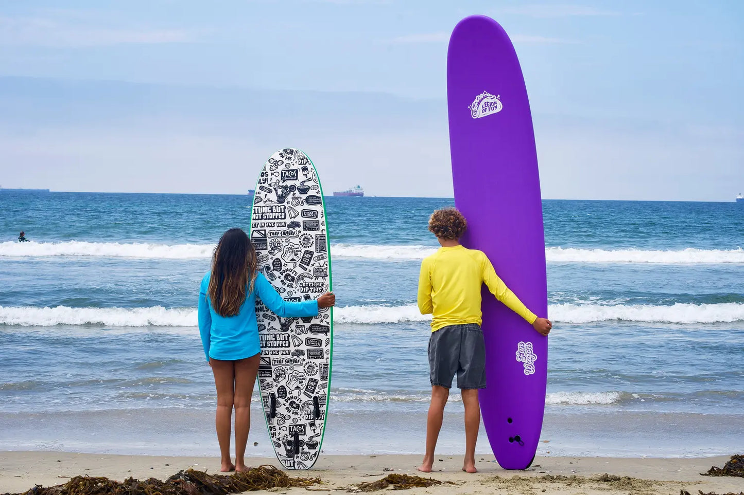 Two teens face the SoCal ocean holding foam surfboards, one 7' surfboard, one 10' surfboard, on the beach