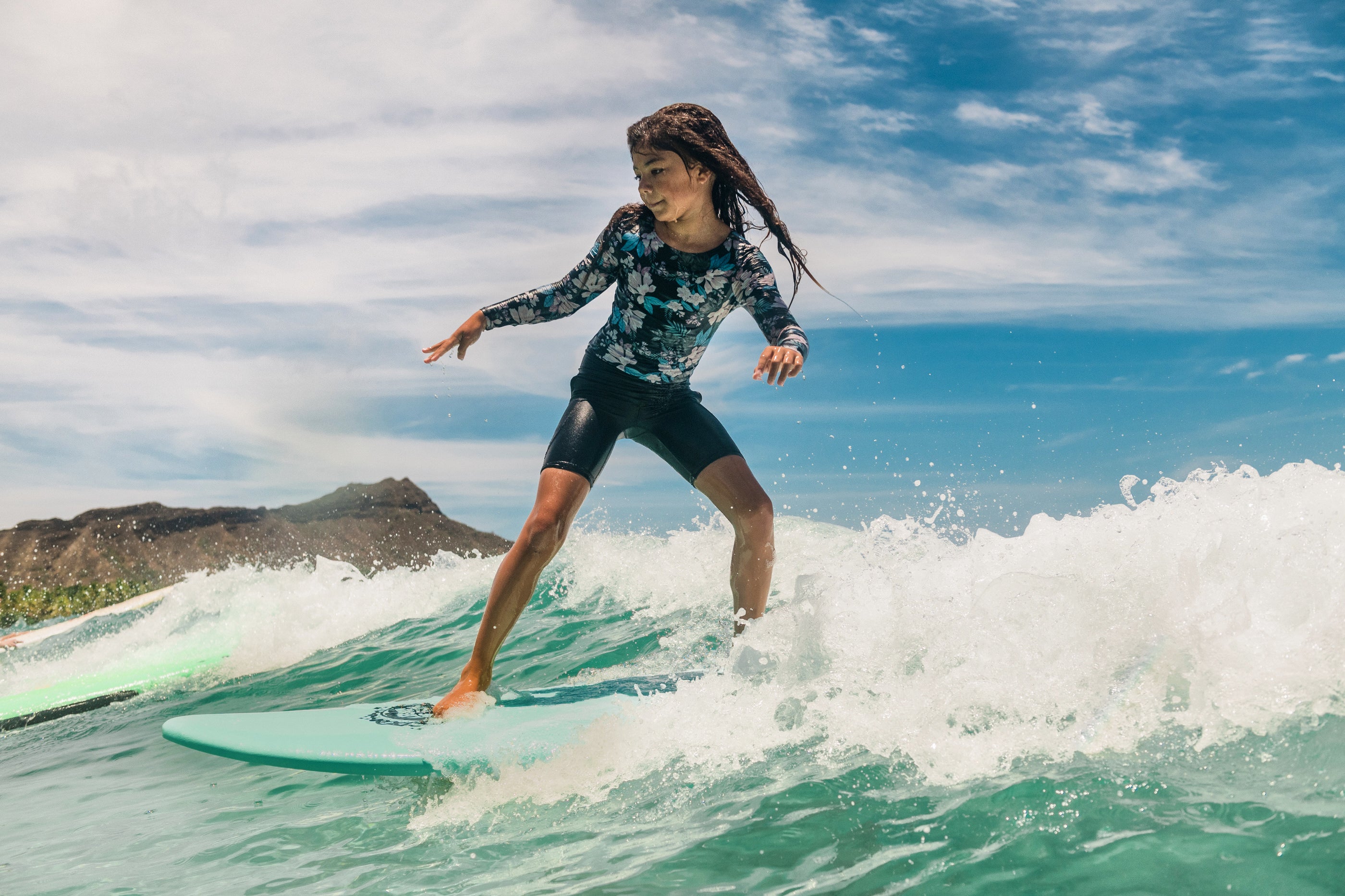 Girl surf riding on a Legion of Fun surfboard, surfing a wave in Hawaii with a mountain and blue sky in the background