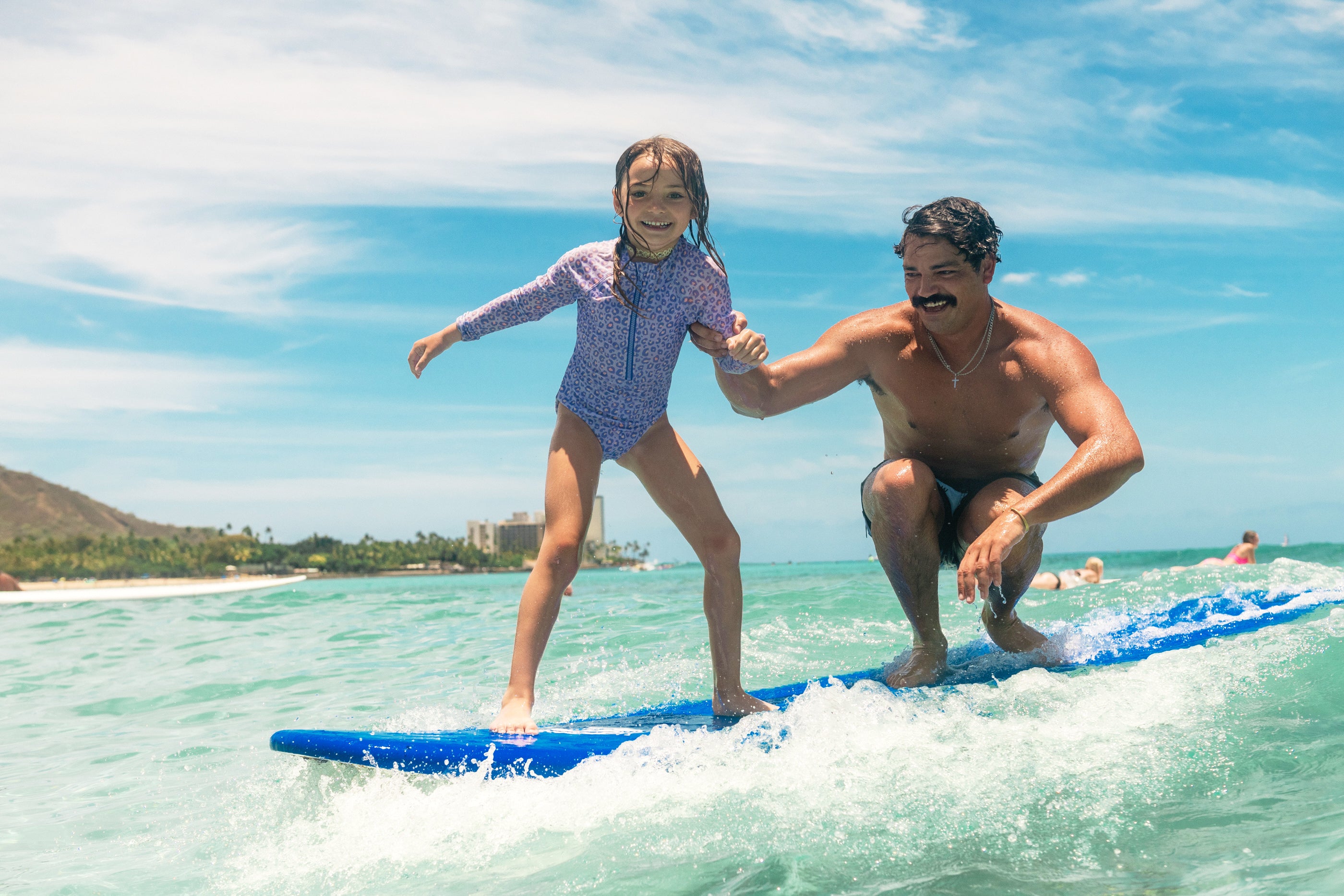 Father and daughter surfing together on a blue Legion of Fun surfboard in clear water with a blue sky.